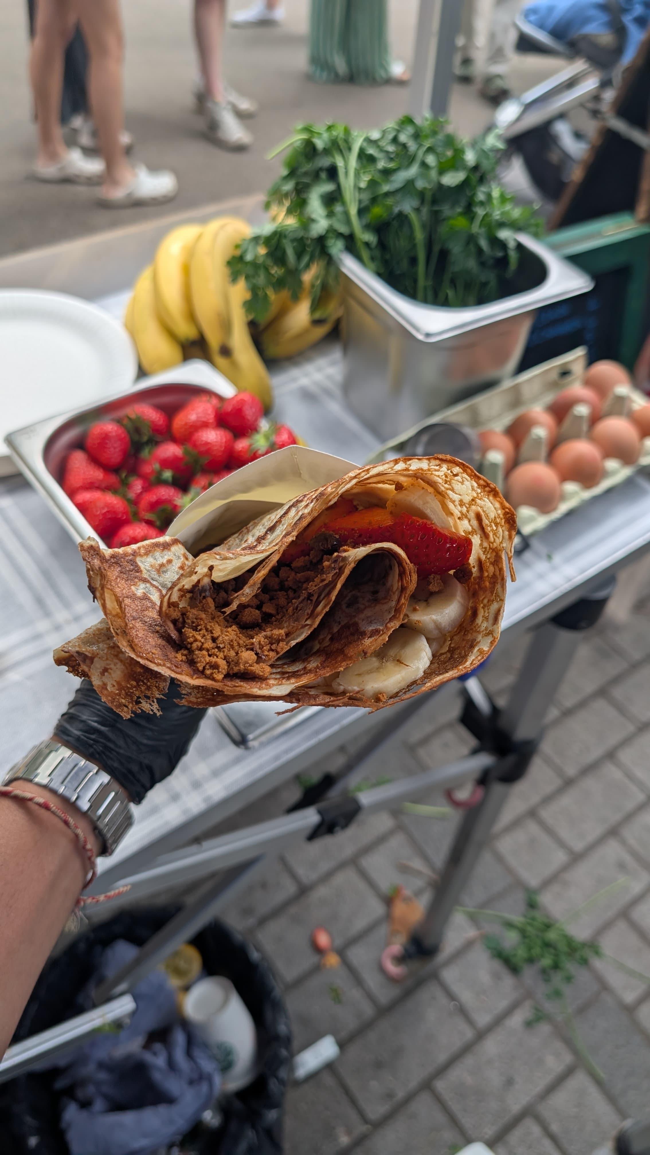 Hand-held sweet crêpe wrap with strawberries, banana and speculoos — gloved hand, event in the background